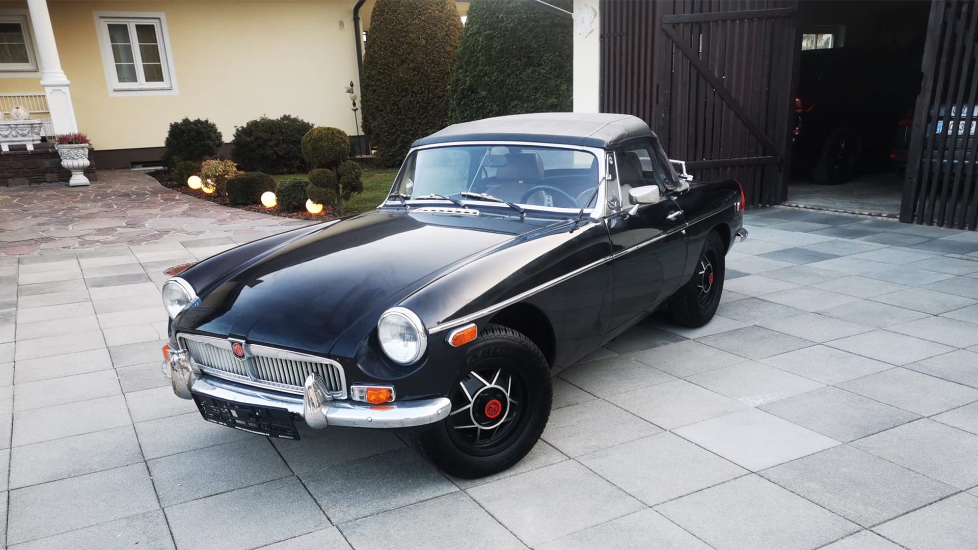Schwarzer MG B Roadster mit geschlossenem Verdeck in der Abendsonne vor der Werkstätte